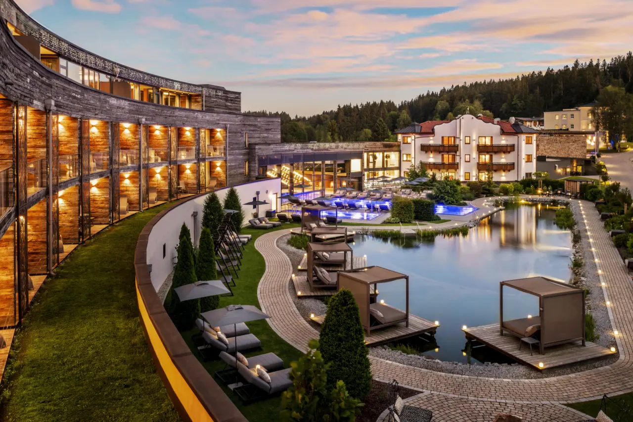 Wellnessresort aus der Vogelperspektive in der Abenddämmerung, geschwungener Holzbau mit beleuchteten Balkonen, Badeteich mit Stegen und beheizte Außenpools