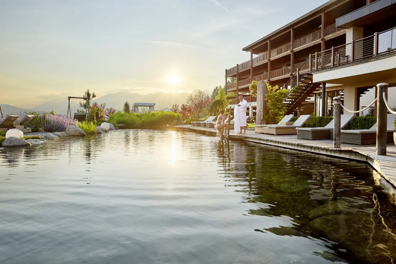 Hotel Chalet Mirabell in Hafling – Naturbadeteich mit Gästen und Hotelgebäude im warmen Abendlicht | Wellnesshotel Südtirol
