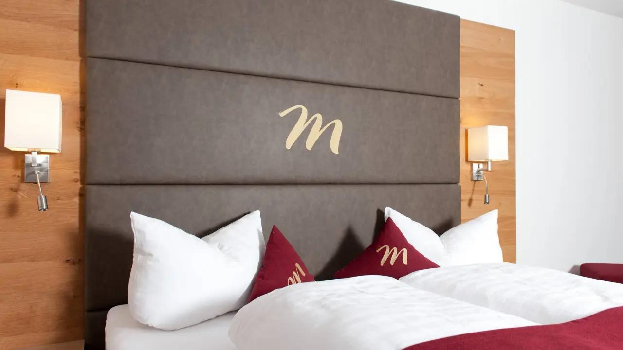 Hotel Mallaun in See/Paznaun – Komfortables Doppelbett mit gepolstertem Kopfteil und Logo | Wellnesshotel Paznaun