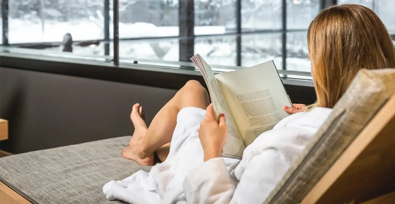 TRAUBE BRAZ Alpen.Spa.Golf.Hotel in Braz – Frau entspannt im Bademantel mit Buch und Winterblick | Wellnesshotel Vorarlberg