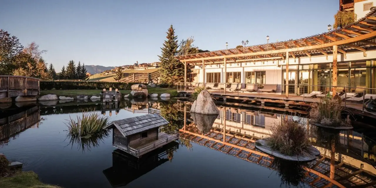 Hotel Bacherhof in Terenten – Naturbadeteich mit Holzsteg und Hotelgebäude | Wellnesshotel Südtirol